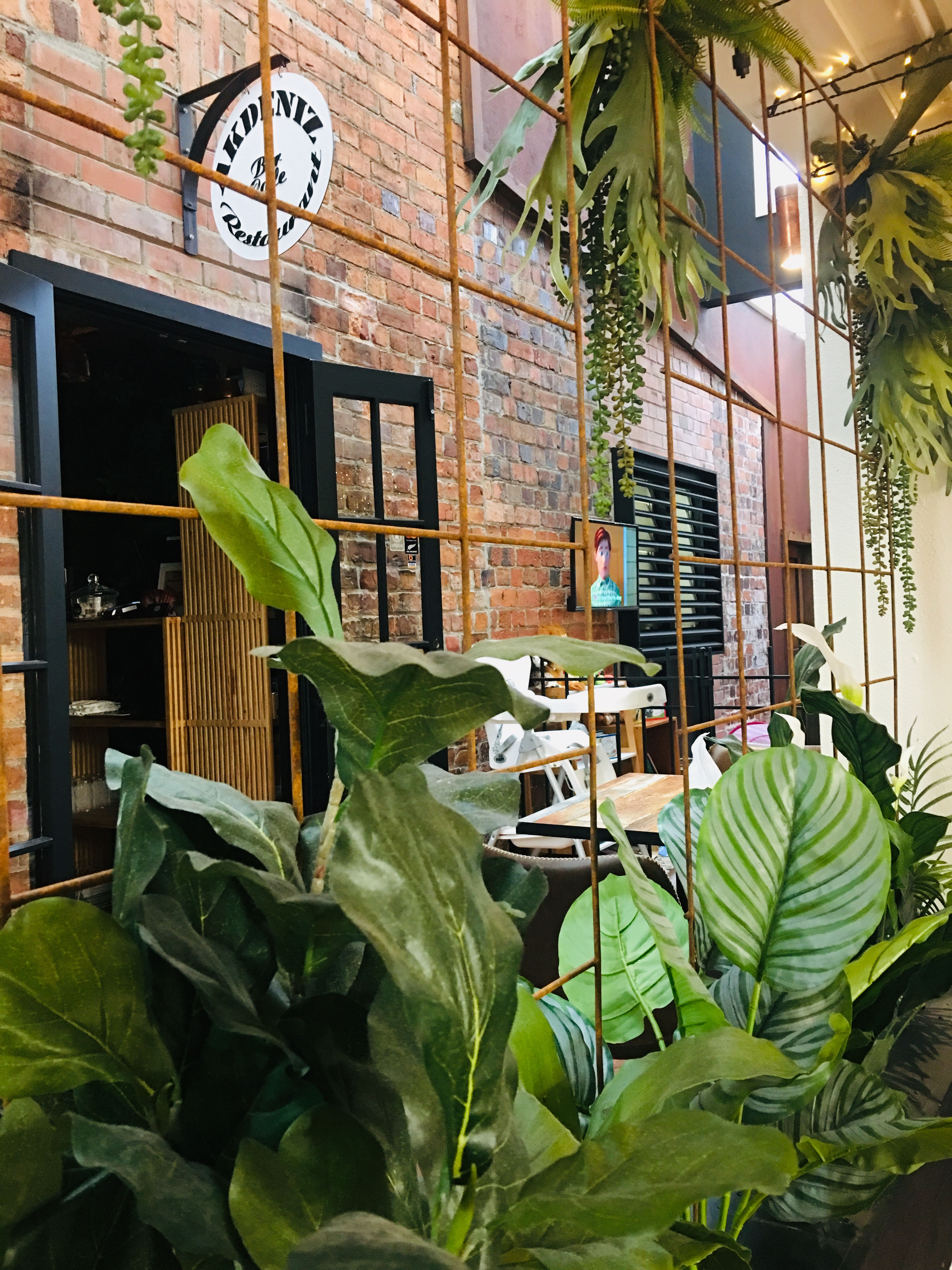 The entrance to Akdeniz seen through lush green plants — the round AKDENIZ signboard on the red-brick wall of the Devonport Arcade